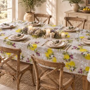 Tovaglia beige con limoni e olive su tavolo per sei, con piatti crema, calici, candele e fiori secchi.