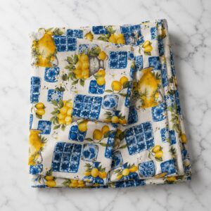 Tovaglia limone e maiolica siciliana cotone piegata su marmo bianco – fantasia limoni gialli dorati, piastrelle blu e bianche e vasi in pietra su fondo crema